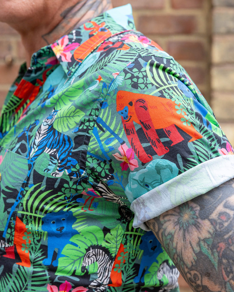 Close up shot of James wearing Jungle Stretch Short Sleeve Shirt. The collared shirt is a forest green colour with all over print of various jungle animals and plants in black, white, red, pink, yellow and blue. Close up shot shows close up of print and rolled up shirt sleeve.