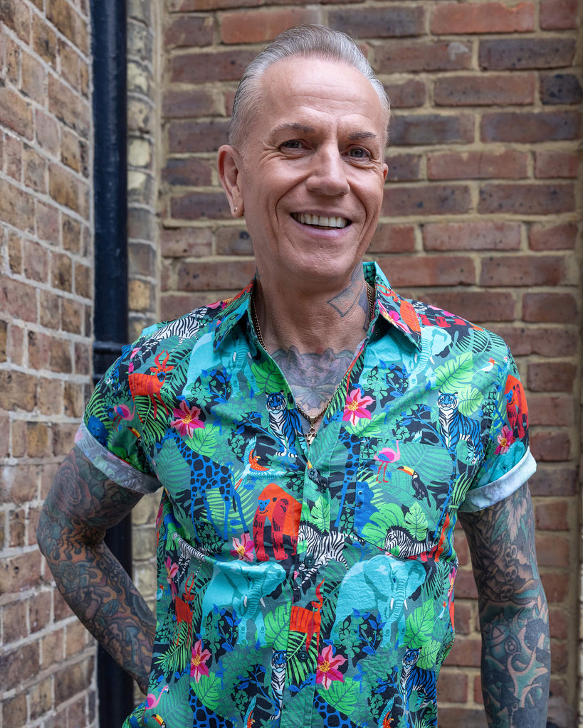 James is wearing Jungle Stretch Short Sleeve Shirt. The collared shirt is a forest green colour with all over print of various jungle animals and plants in black, white, red, pink, yellow and blue. James is smiling at the camera with one hand on his hip.