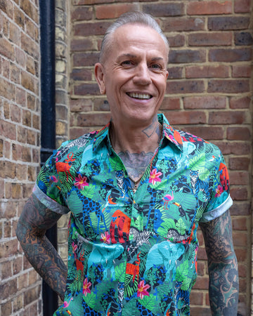 James is wearing Jungle Stretch Short Sleeve Shirt. The collared shirt is a forest green colour with all over print of various jungle animals and plants in black, white, red, pink, yellow and blue. James is smiling at the camera with one hand on his hip.