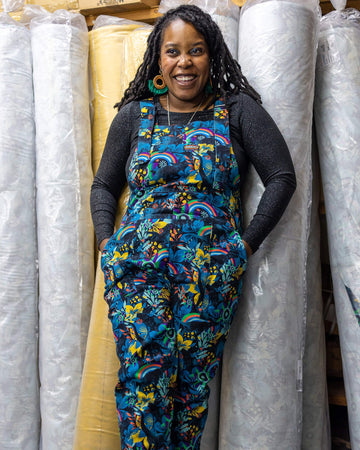 Ama is standing in a warehouse next to rolls of fabric, smiling with hands in pockets wearing the black jurassic stretch twill dungarees with a black long sleeve top underneath and brown boots. 