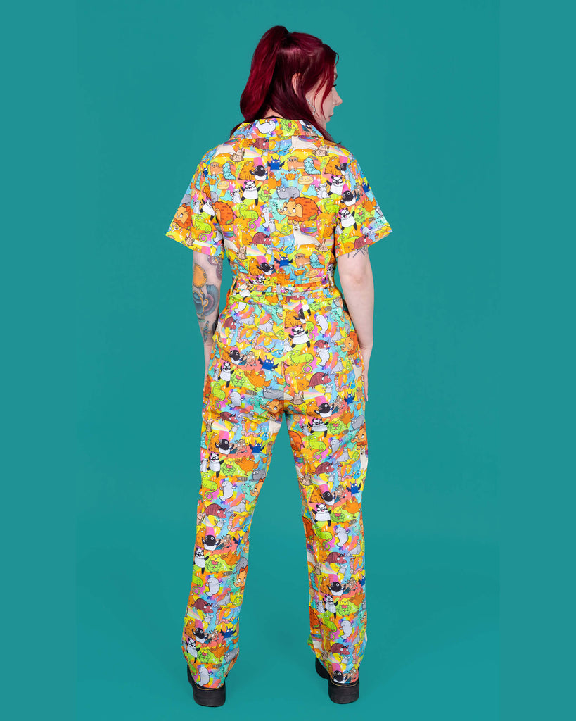Florence a red haired female model with tattoos and cool make up is wearing a colourful rainbow jumpsuit with cute drawing by Katie Abey. She is standing against a turquoise background ands her back to camera 