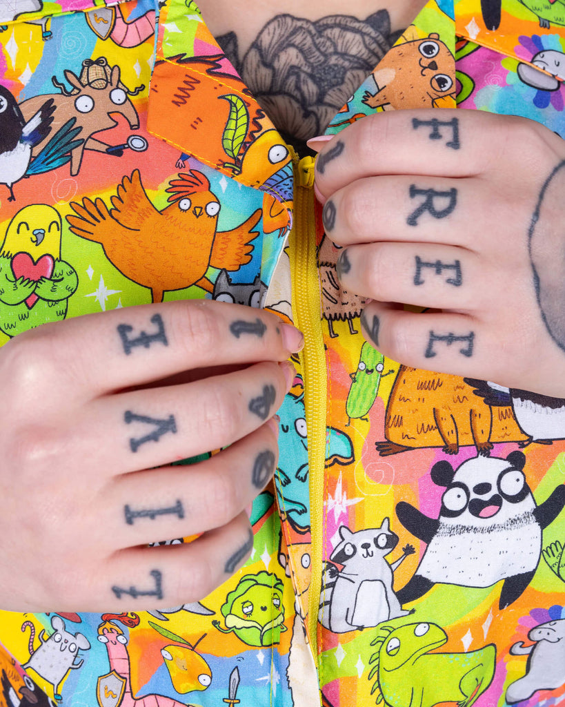 Florence a red haired female model with tattoos and cool make up is wearing a colourful rainbow jumpsuit with cute drawing by Katie Abey. She is standing against a turquoise background. This is a close up of her tattooed hands on the zip