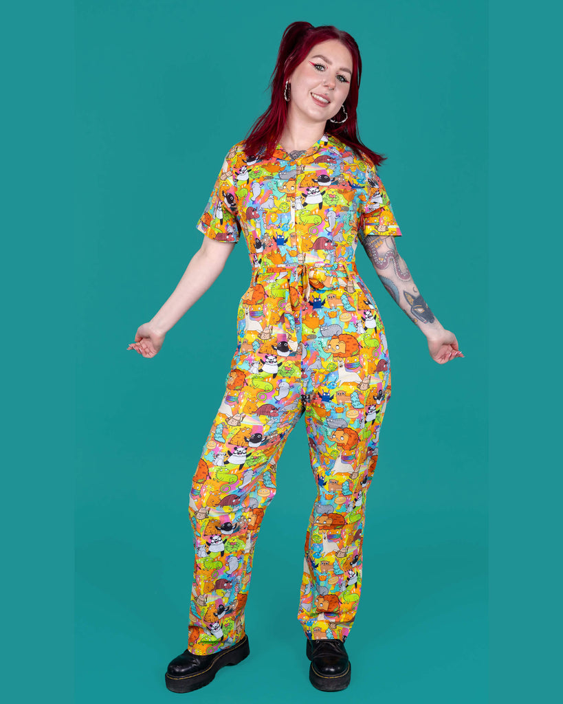 Florence a red haired female model with tattoos and cool make up is wearing a colourful rainbow jumpsuit with cute drawing by Katie Abey. She is standing against a turquoise background and smiling at camera. 