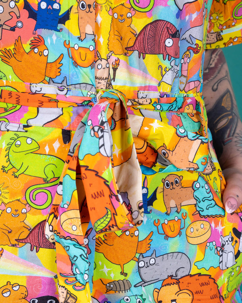 Florence a red haired female model with tattoos and cool make up is wearing a colourful rainbow jumpsuit with cute drawing by Katie Abey. She is standing against a turquoise background this is a close up of the tie belt fastening