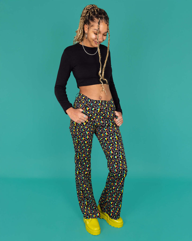 Milly a femme model with blonde locs is wearing Run & Fly Grey Rainbow Leopard Print Bell Bottom Flares paired with a long sleeve black crop top and green boots. The flares are a dark grey with an all over rainbow leopard print. Milly is looking down and smiling with her hands in the pockets of the jeans in front of a green background.