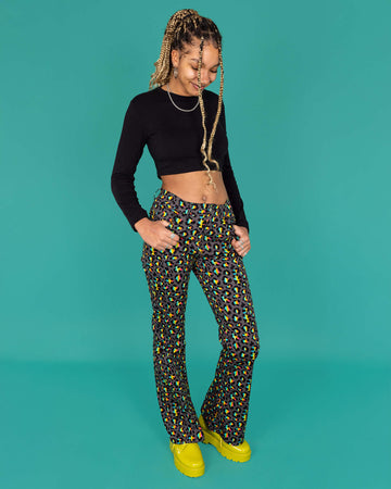 Milly a femme model with blonde locs is wearing Run & Fly Grey Rainbow Leopard Print Bell Bottom Flares paired with a long sleeve black crop top and green boots. The flares are a dark grey with an all over rainbow leopard print. Milly is looking down and smiling with her hands in the pockets of the jeans in front of a green background.