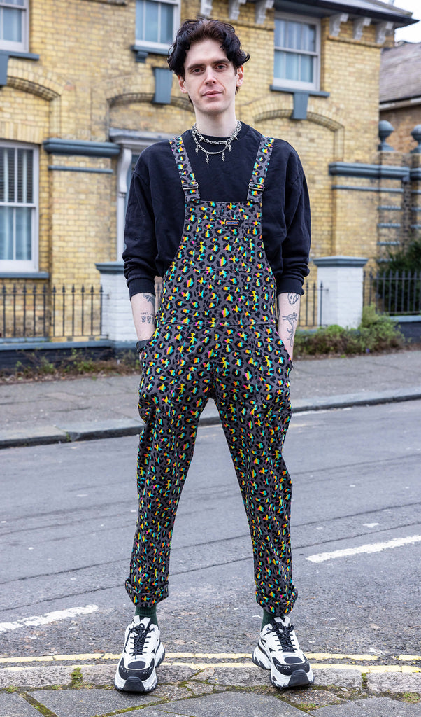 Jake a white male with dark hair is wearing Run & Fly Grey Rainbow Leopard Stretch Twill Dungarees paired with a black sweatshirt and white and black trainers. The grey dungarees have an all over rainbow leopard print design. Jake is stood facing the camera with his hands in the dungaree pockets outdoors in Hove. 