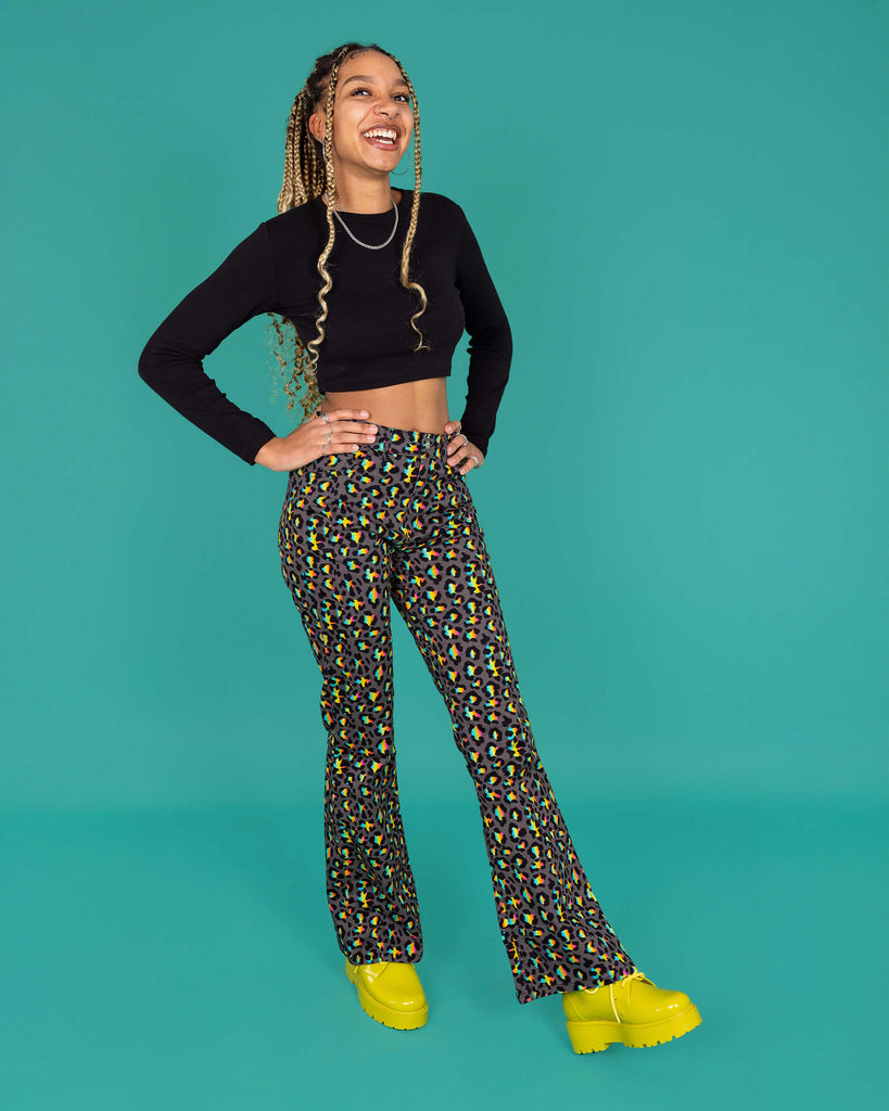 Milly a femme model with blonde locs is wearing Run & Fly Grey Rainbow Leopard Print Bell Bottom Flares paired with a long sleeve black crop top and green boots. The flares are a dark grey with an all over rainbow leopard print. Milly is smiling with her hands on her hips and one leg in front of the other in front of a green background. 