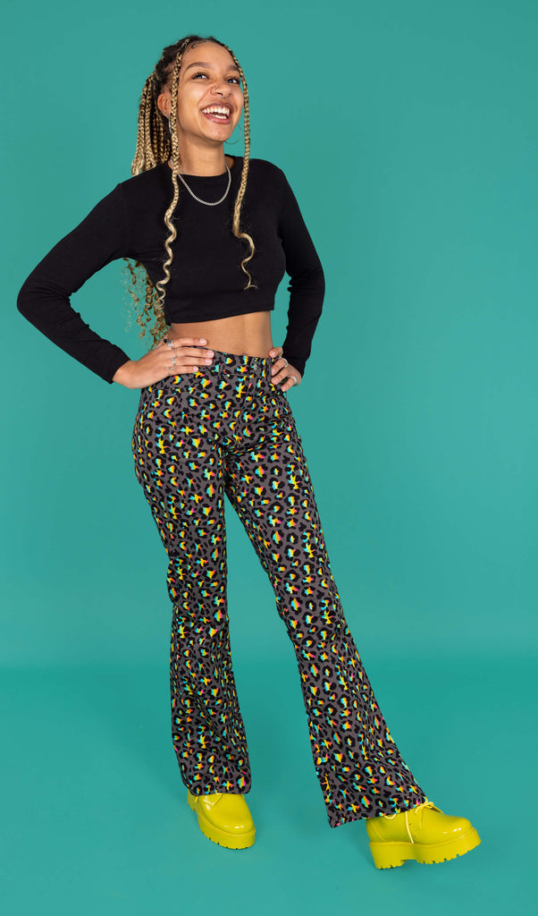 Milly a femme model with blonde locs is wearing Run & Fly Grey Rainbow Leopard Print Bell Bottom Flares paired with a long sleeve black crop top and green boots. The flares are a dark grey with an all over rainbow leopard print. Milly is smiling with her hands on her hips and one leg in front of the other in front of a green background. 