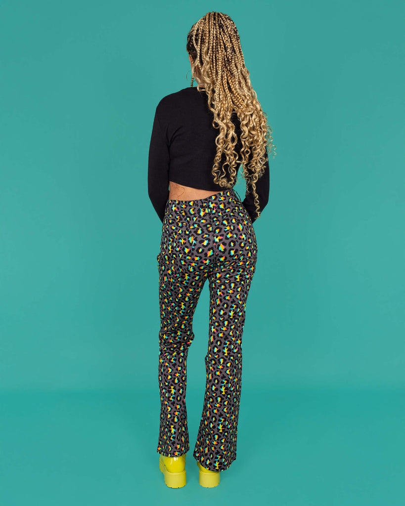 Milly a femme model with blonde locs is wearing Run & Fly Grey Rainbow Leopard Print Bell Bottom Flares paired with a long sleeve black crop top and green boots. The flares are a dark grey with an all over rainbow leopard print. Milly is stood with her back to the camera with one leg bent in front of a green background. 
