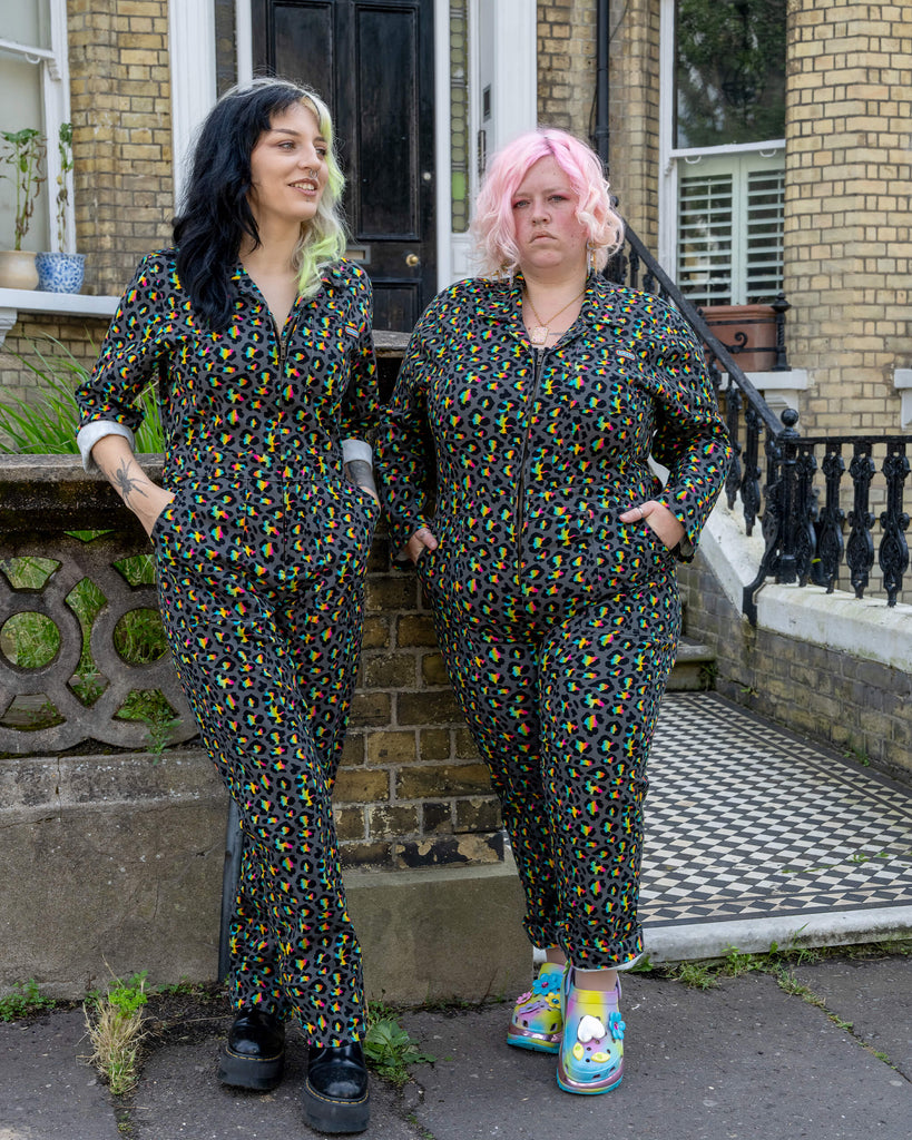 Two models, one with black and green hair and the other with pink hair, stand side by side in the Rainbow Leopard Stretch Twill Boiler Suit by Run & Fly. The bright, multicoloured leopard print contrasts against the brick backdrop of a townhouse. The models embody the playful spirit of the ethical slow fashion brand, showcasing bold and creative style choices.