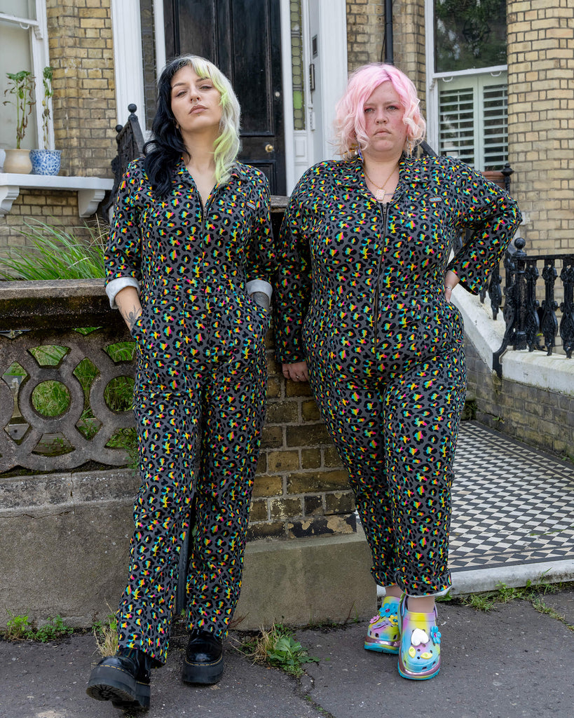 Two models stand side by side wearing the Rainbow Leopard Stretch Twill Boiler Suit by Run & Fly. One model has black and green hair, the other has pink hair. Both wear the brightly coloured, multicoloured leopard print designs, contrasting with the urban Victorian-style setting. Run & Fly’s ethical slow fashion pieces are designed to stand out.
