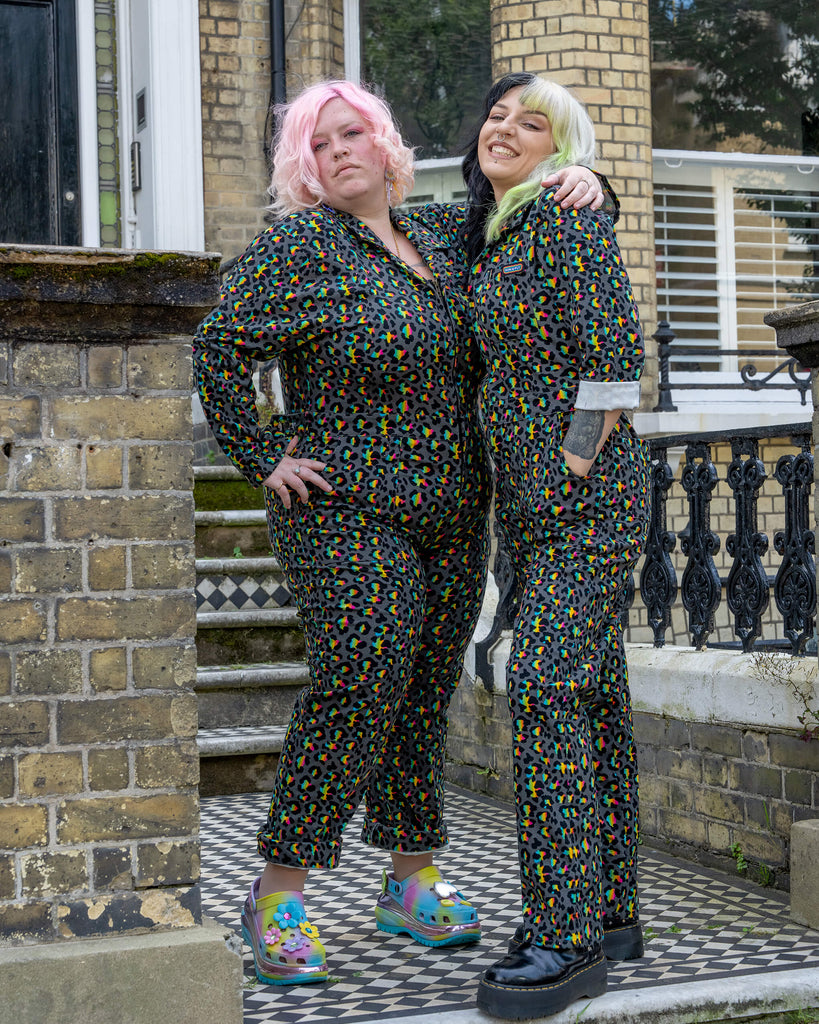 Two models wearing the Rainbow Leopard Stretch Twill Boiler Suit by Run & Fly pose in front of a townhouse. The models, with pink and black/green hair, showcase the suit’s fun and vibrant leopard print design. The bold colours and confident styling embody the playful spirit of this ethical slow fashion brand.