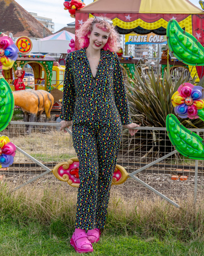 A young woman with pink curly hair, wearing a stylish black jumpsuit with a vibrant neon leopard print pattern, stands smiling at an outdoor amusement park. She accessorises with black sunglasses and bright pink Crocs over pink socks. The background features colourful carnival rides and decor, including a mini-golf sign and whimsical animal figures.