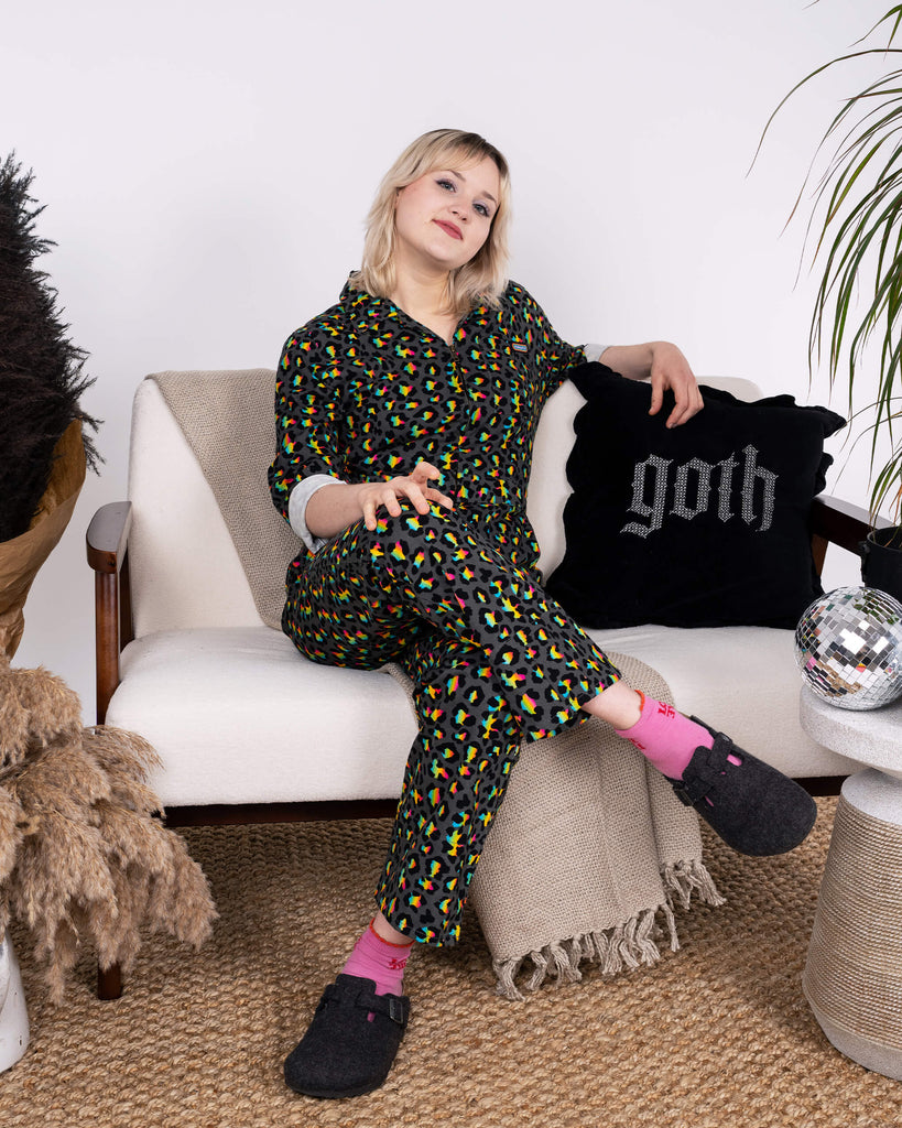 The model relaxes on a cosy chair wearing the Rainbow Leopard Stretch Twill Boiler Suit by Run & Fly. Styled with pink socks and slippers, the bold rainbow leopard print stands out in a modern lounge filled with plants and disco balls.