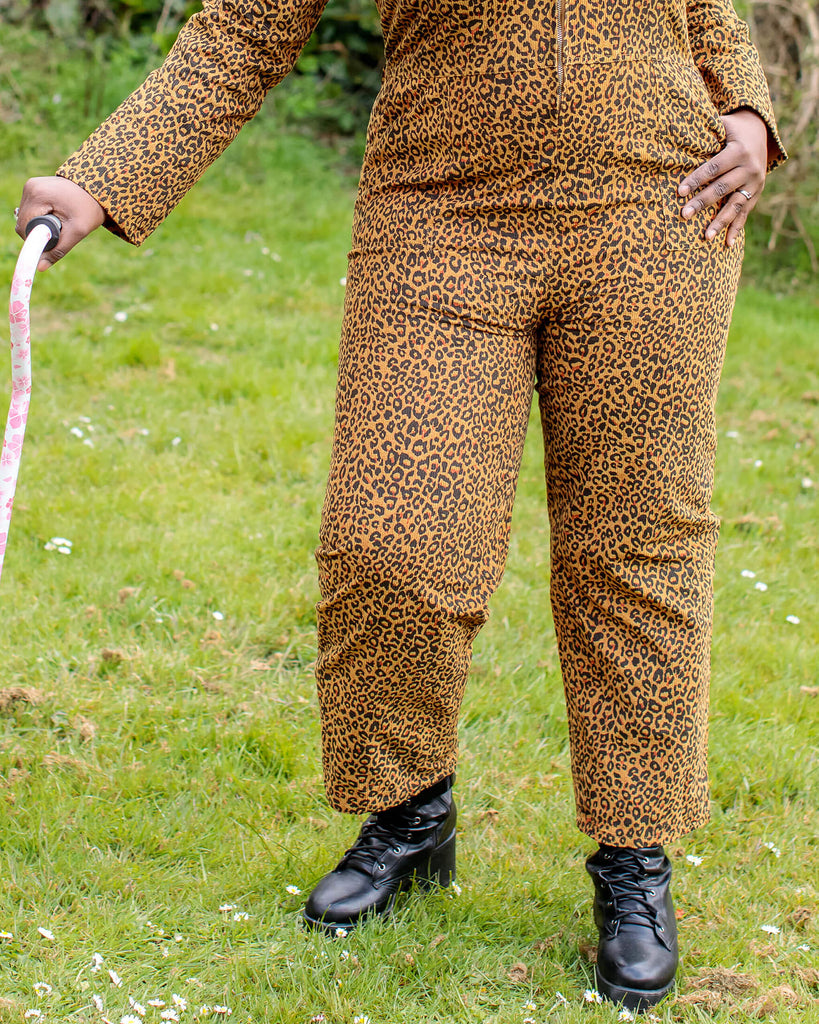 Close up of the legs of Ayesha stood in a garden area wearing the natural leopard print corduroy boiler suit with a black cat necklace and black boots with a pastel pink walking stick. She is posing with one hand in her pocket and looking off to the left smiling.