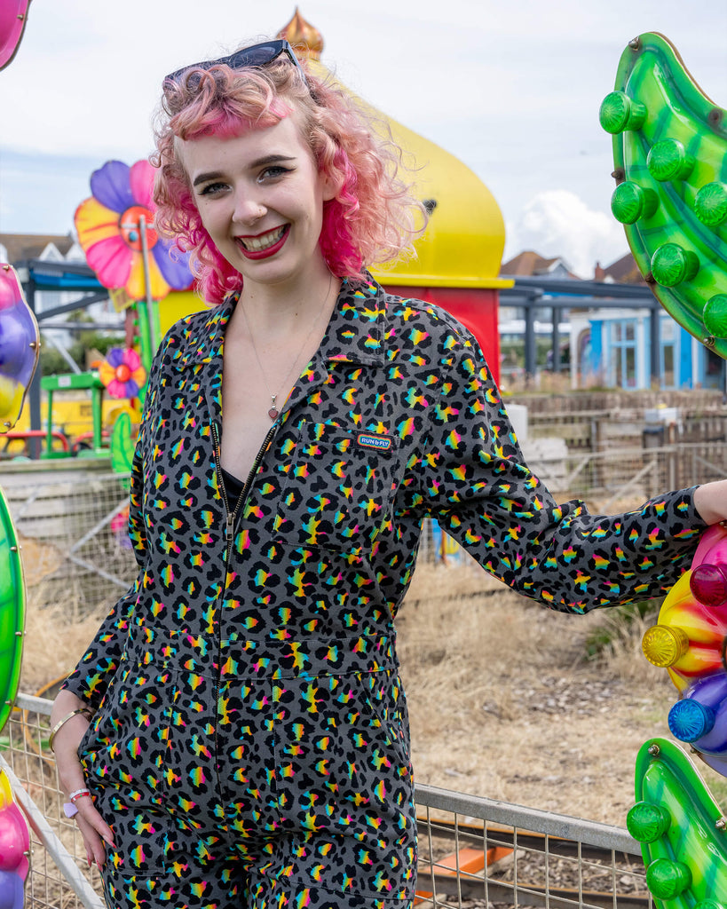 Close-up of a smiling model with curly pink hair wearing a Rainbow Leopard Stretch Twill Boiler Suit by Run & Fly. The suit’s colourful leopard print stands out against the model’s black underlayer. Set in a carnival backdrop, the bright and fun environment highlights the vibrant, slow-fashion brand’s creative design.