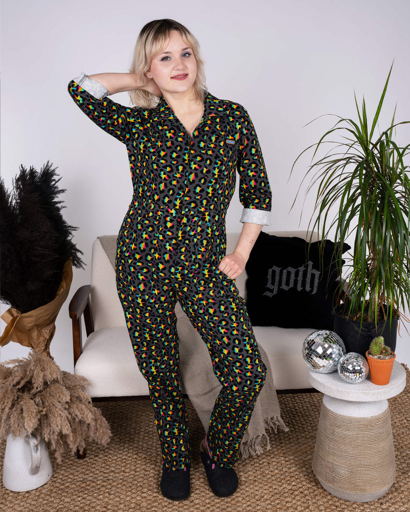 A model poses confidently in the Rainbow Leopard Stretch Twill Boiler Suit by Run & Fly, a small ethical slow fashion brand. The black suit features a vibrant rainbow leopard print with a relaxed fit and turned-up cuffs, styled in a cosy lounge setting with plants and gothic-themed decor.