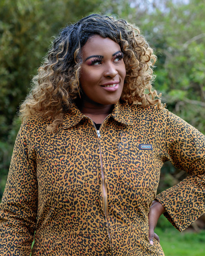 Ayesha is stood in a garden area wearing the natural leopard print corduroy boiler suit with a black cat necklace and black boots with a pastel pink walking stick. She is posing with one hand on her hip smiling looking off to the right. Photo is cropped from the hips up.