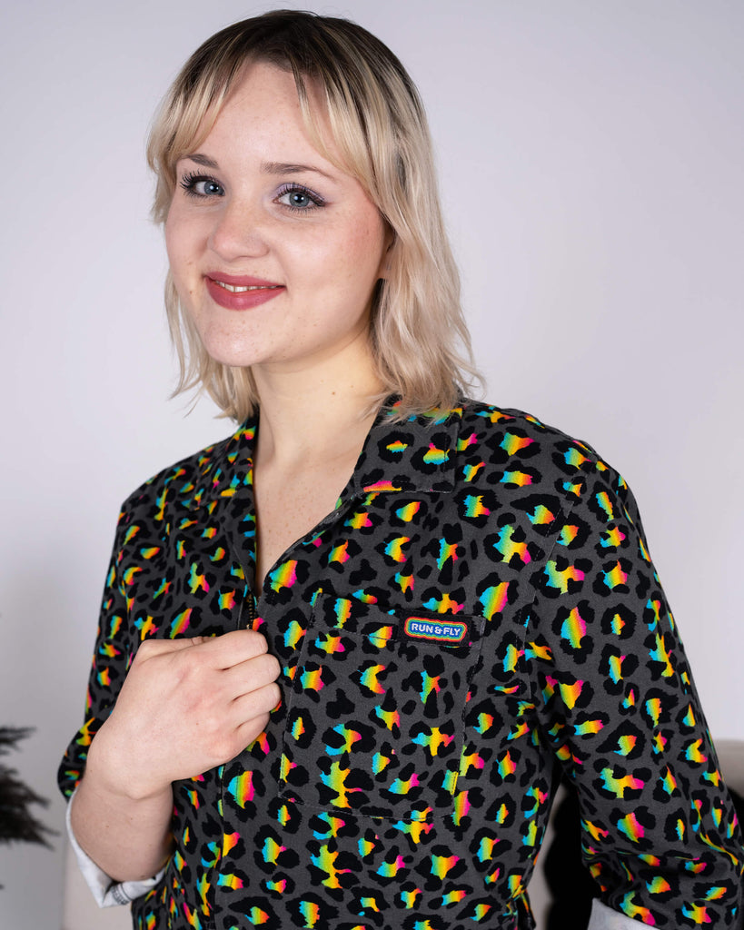 Close-up of the Rainbow Leopard Stretch Twill Boiler Suit by Run & Fly, highlighting its bright rainbow leopard print on a black base. The model smiles while showcasing the brand's logo on the chest pocket. Ethical slow fashion at its boldest and most playful.