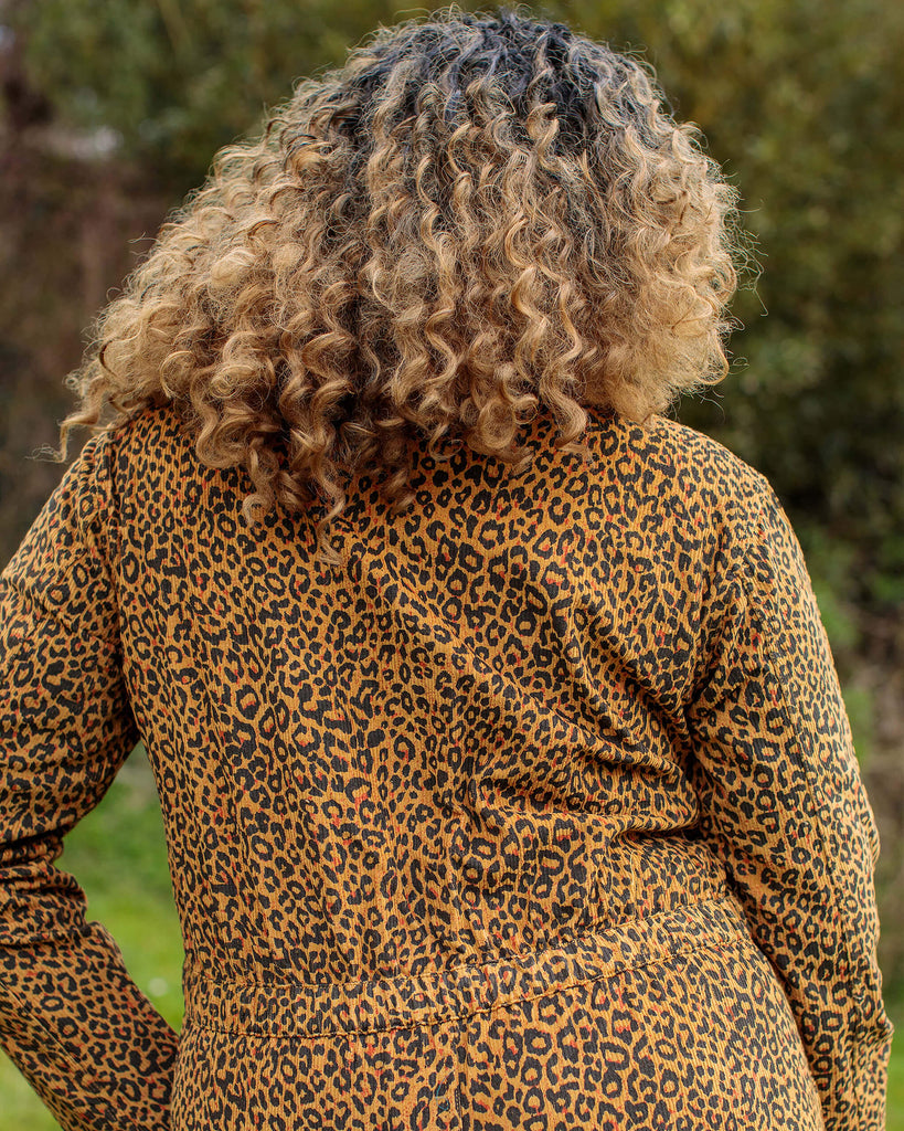 Ayesha is stood in a garden area wearing the natural leopard print corduroy boiler suit with a black cat necklace and black boots with a pastel pink walking stick. She is facing away from the camera with one hand in the front pocket.