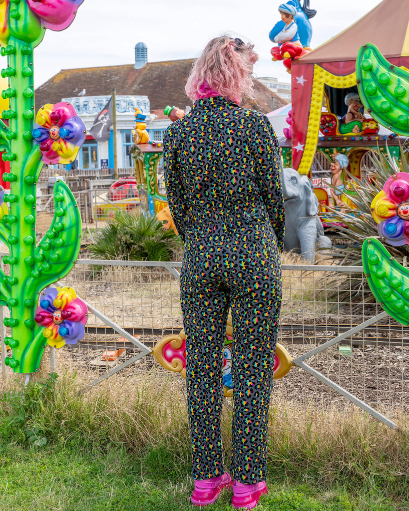 Back view of a woman with pink curly hair, wearing a black jumpsuit with a neon leopard print pattern. She stands at an outdoor amusement park, with colourful carnival rides in the background, including an elephant figure and bright decor. The outfit highlights the playful yet stylish design suitable for fun outdoor activities.