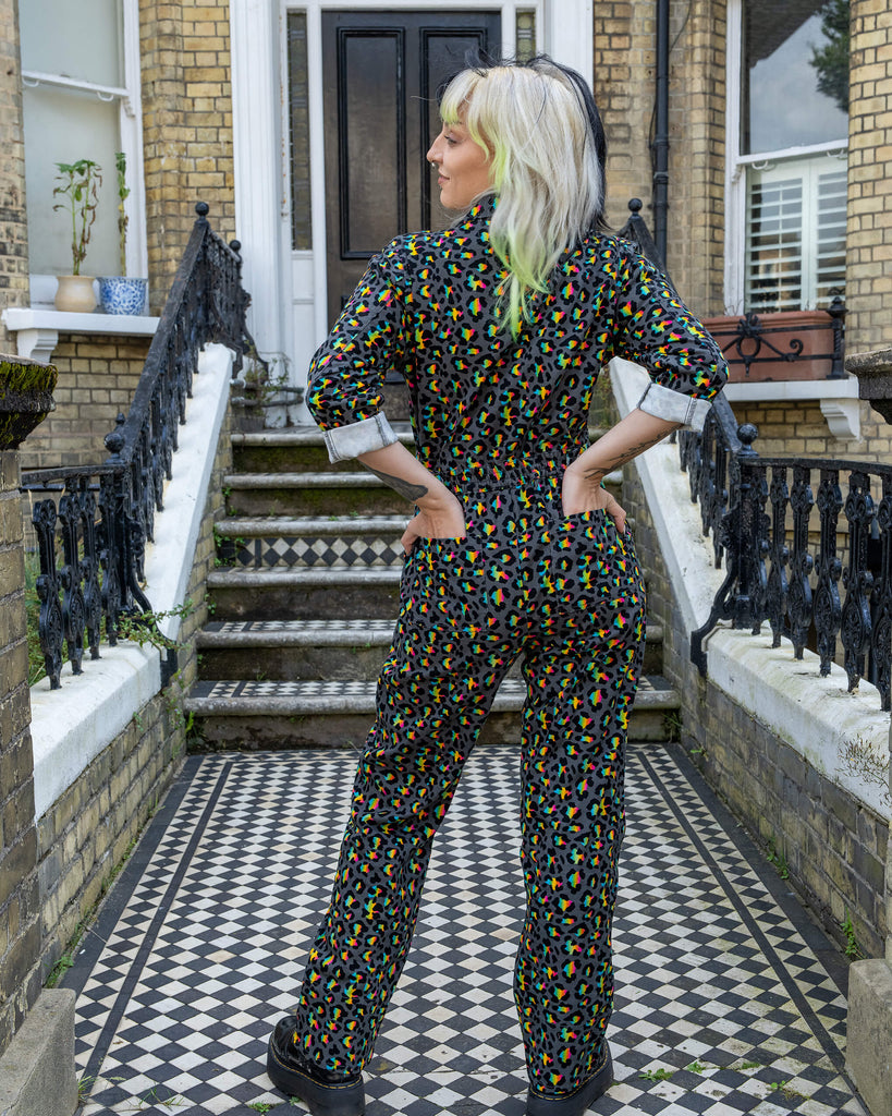 The same model with black and green hair showcases the back view of the Rainbow Leopard Stretch Twill Boiler Suit by Run & Fly. The suit fits snugly around the waist with an elasticated back, displaying the playful multicolour leopard print in full. Set against a black-and-white tiled doorstep, the contrast highlights the boldness of the outfit.