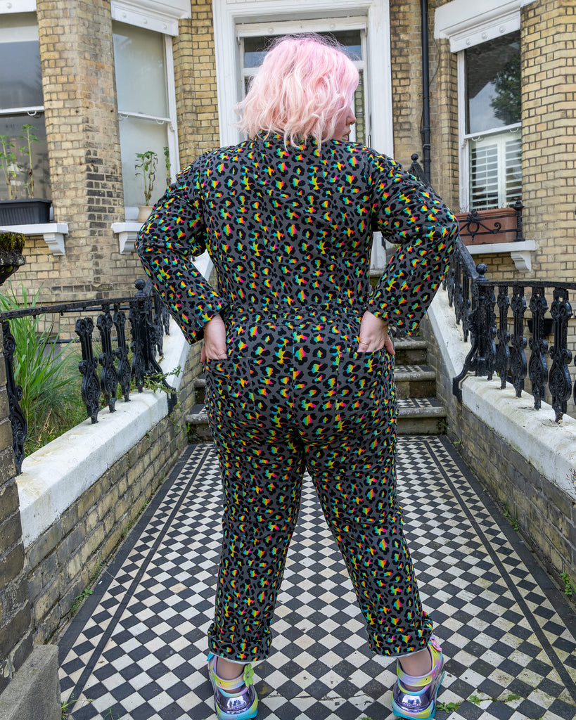 Back view of a model with pink hair wearing the Rainbow Leopard Stretch Twill Boiler Suit by Run & Fly. The multicoloured leopard print contrasts with the Victorian-style home backdrop, showcasing the playful design of this ethical slow fashion piece. The model’s hands rest on her hips, highlighting the suit's comfortable, relaxed fit.