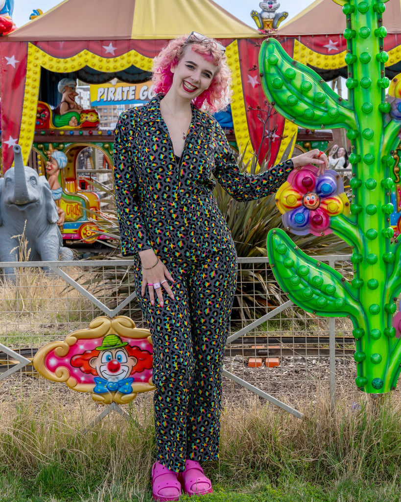 A model with curly pink hair wears a Rainbow Leopard Stretch Twill Boiler Suit by Run & Fly, standing next to a bright carnival display. The suit features a vibrant multicolour leopard print on a black background, adding a fun pop of colour. The small ethical fashion brand creates bold and unique designs. The model's wide smile and pink Crocs complete the playful look.