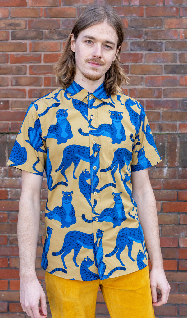 Jack is stood in front of a brick wall wearing the blue leopard short sleeve shirt with gold corduroy flares. They are facing forward posing with both hands resting by their sides and smiling whilst looking to camera. Photo is cropped from the hips up.