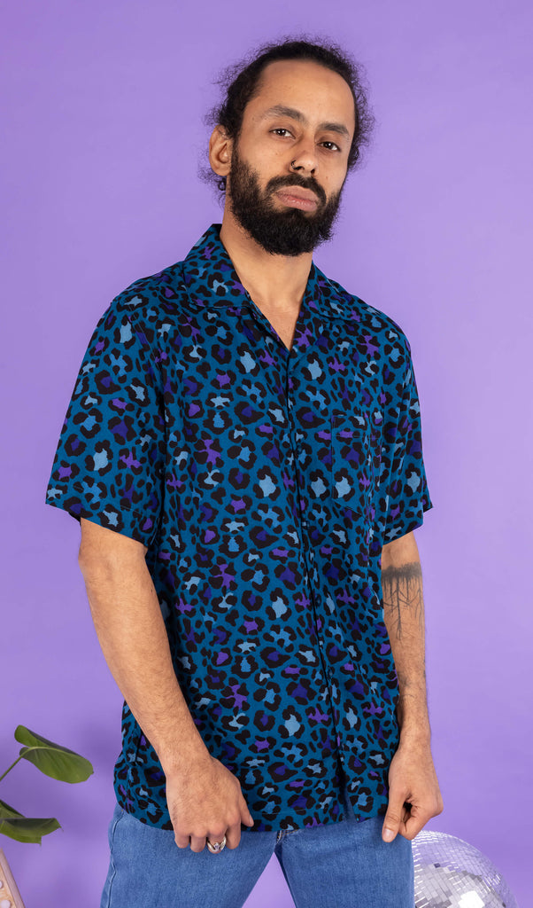 Richard, a hispanic male model with dark hair in a bun and a beard, is stood in a photography studio in Hove in front of a lilac backdrop wearing Atlantic Blue Leopard Print Short Sleeve Rayon Shirt with blue jeans. Richard is posing to the camera with his hands resting at the bottom of the shirt. The photo is cropped at the thighs.