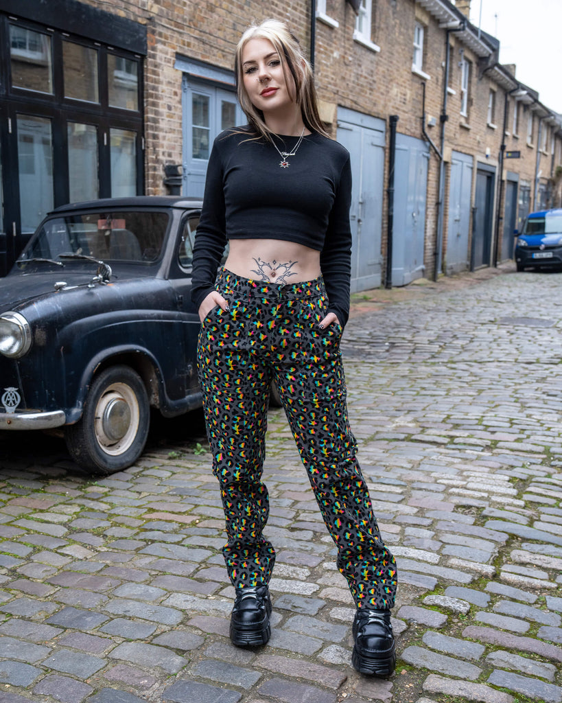 Front view of a smiling model wearing Run & Fly Grey Rainbow Leopard Straight Leg Trousers, hands in pockets, styled with a black long-sleeve crop top and chunky black shoes, standing on a cobbled street.