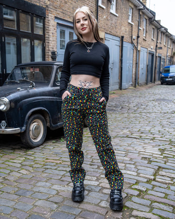 Front view of a smiling model wearing Run & Fly Grey Rainbow Leopard Straight Leg Trousers, hands in pockets, styled with a black long-sleeve crop top and chunky black shoes, standing on a cobbled street.