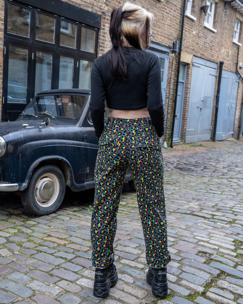 Back view of a model wearing Run & Fly Grey Rainbow Leopard Straight Leg Trousers, standing on a cobbled street with a vintage black car in the background. The trousers feature a grey base with vibrant rainbow leopard print and are styled with black platform boots and a cropped long-sleeve top.