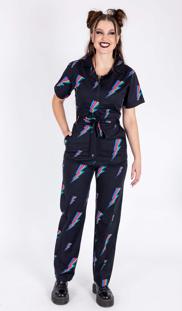 A model is stood in a white background wearing the Run & Fly new and improved black rainbow lightning jumpsuit with black patent boots. They are facing forward with one hand in their pocket, other hand by their side and laughing looking right. The jumpsuit is all black with a belt tied into a ribbon with a zip up front and covered in rainbow multicoloured lightning bolts all over. The slow fashion jumpsuit features a 70s style flat collar with short sleeves & wide legs.