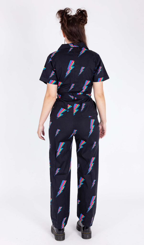 A model is stood in a white background wearing the Run & Fly new and improved black rainbow lightning jumpsuit with black patent boots. They are facing away from the camera to highlight the back detailing. The jumpsuit is all black with a belt tied into a ribbon with a zip up front and covered in rainbow multicoloured lightning bolts all over. The slow fashion jumpsuit features a 70s style flat collar with short sleeves & wide legs.