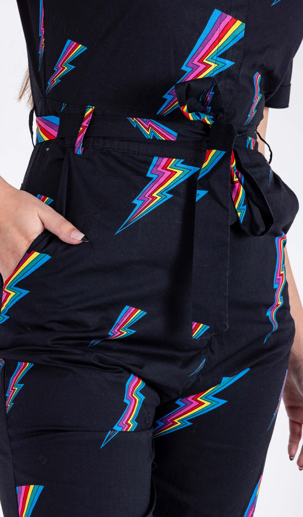 A close up of a model is stood in a white background wearing the Run & Fly new and improved black rainbow lightning jumpsuit. The jumpsuit is all black with a belt tied into a ribbon with a zip up front and covered in rainbow multicoloured lightning bolts all over. The slow fashion jumpsuit features a 70s style flat collar with short sleeves & wide legs.
