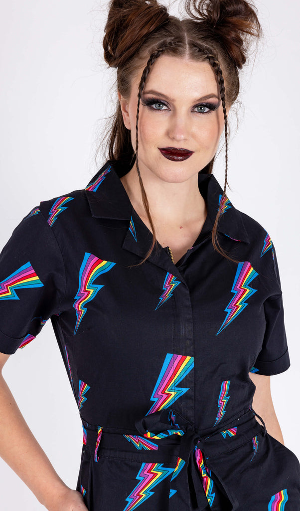 A close up of a model is stood in a white background wearing the Run & Fly new and improved black rainbow lightning jumpsuit. They are facing forward with one hand in their pocket, the other hand by their side and smiling to camera. The jumpsuit is all black with a belt tied into a ribbon with a zip up front and covered in rainbow multicoloured lightning bolts all over. The slow fashion jumpsuit features a 70s style flat collar with short sleeves & wide legs.