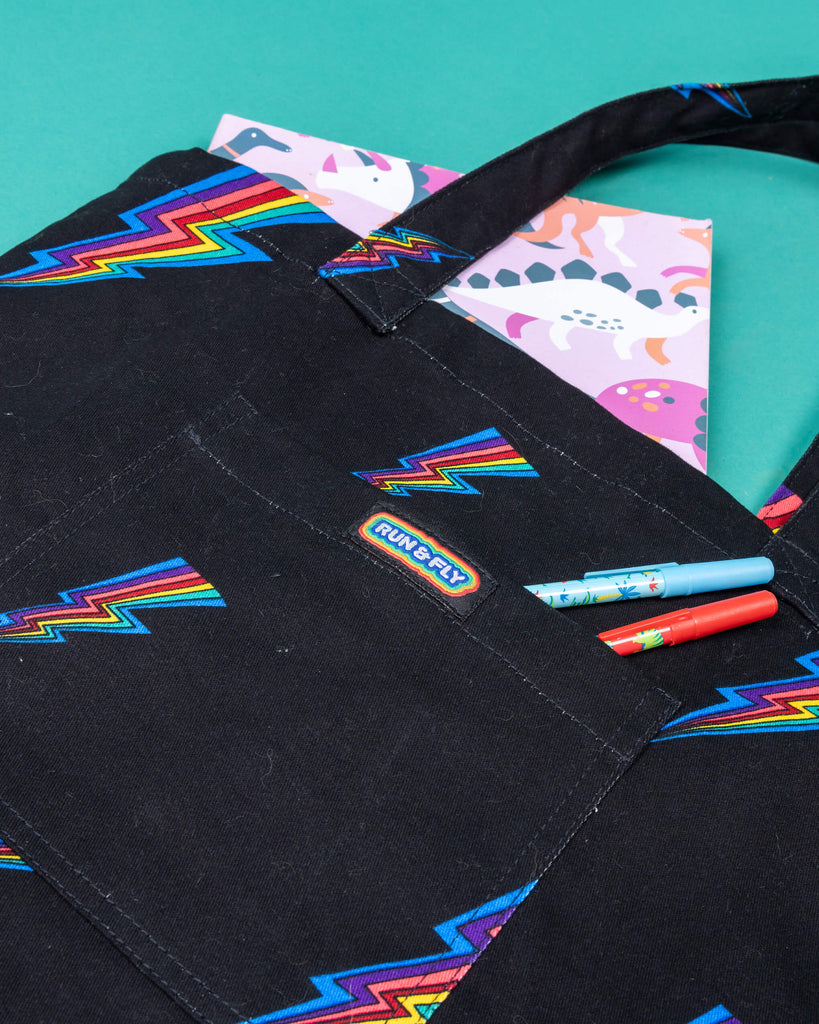 a black tote bag with all over rainbow lightning bolt print. The tote bag is laid flat on a green background with a book in the main section of the bag and two pens in the smaller front pocket with has Run & Fly logo on the front.