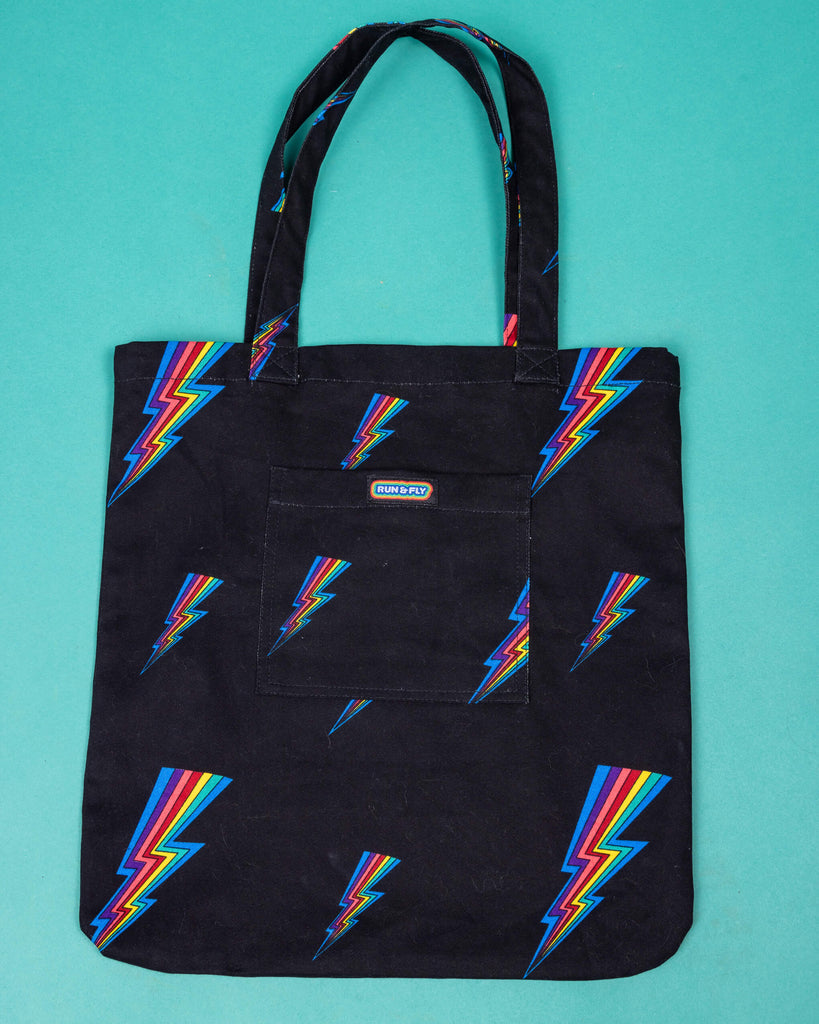 a black tote bag with all over rainbow lightning bold print. The tote bag is laid flat on a green background. The front pocket has Run & Fly logo on the front.