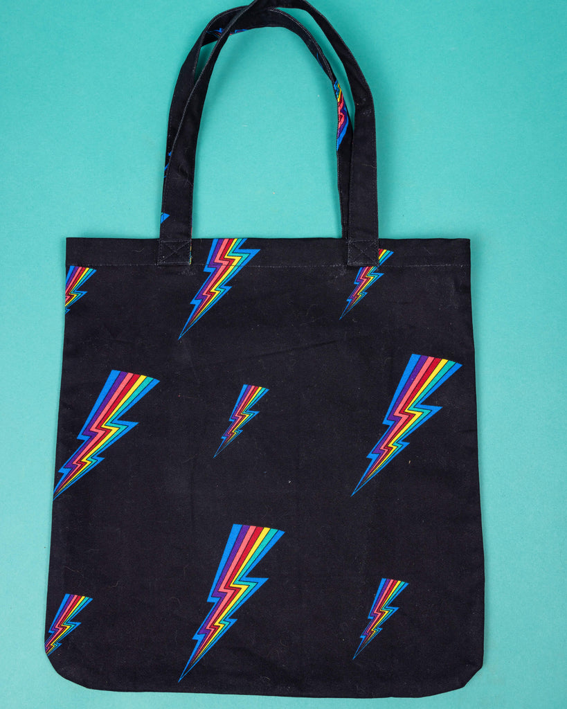 a black tote bag with all over rainbow lightning bold print. The tote bag is laid flat on a green background.