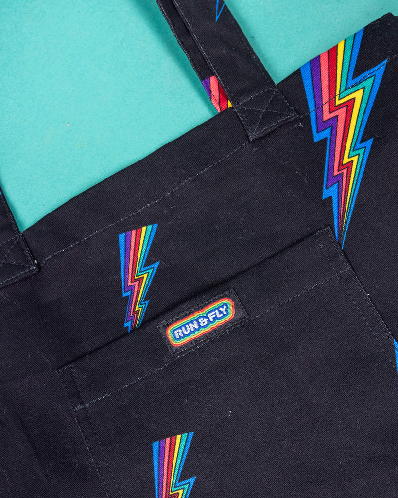 close up of a black tote bag with all over rainbow lightning bold print. The tote bag is laid flat on a green background. The front pocket has Run & Fly logo on the front.