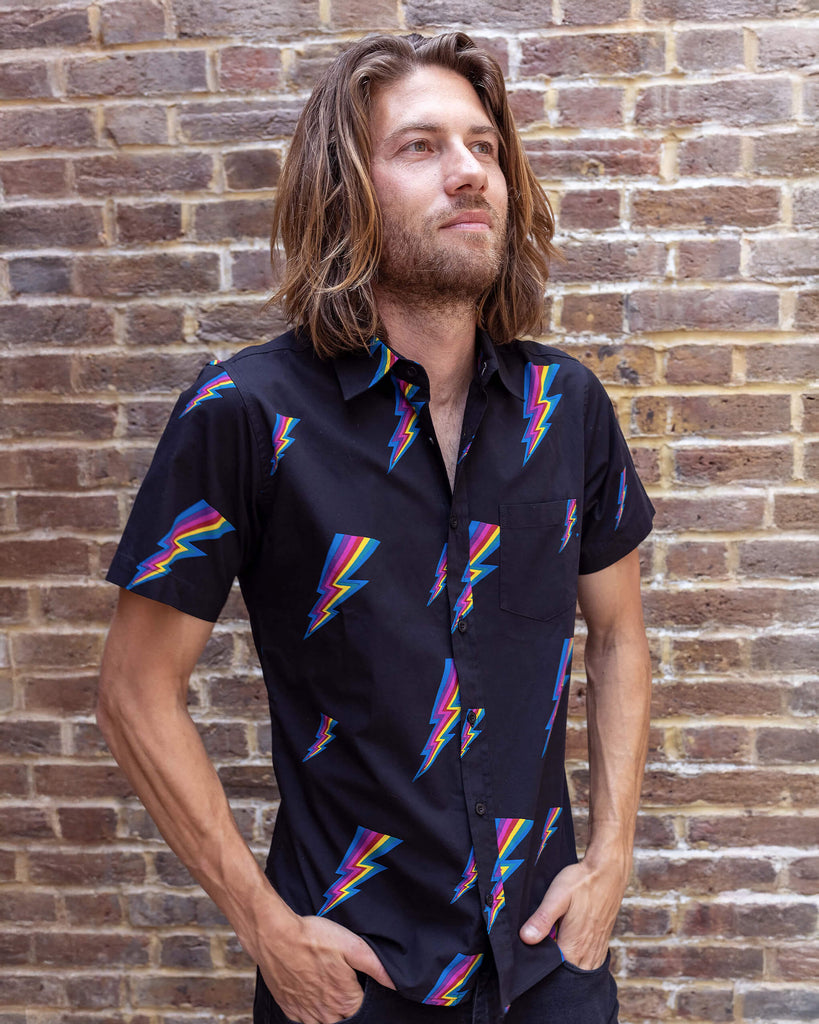 Rudi a male white tanned model with brown hair and a surfer style is wearing a black shirt with lightning bolt design against a brick wall.
