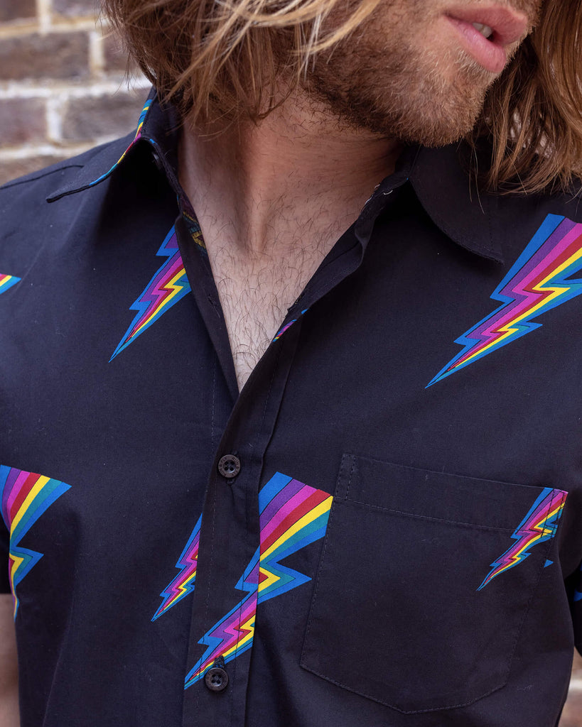 Rudi a male white tanned model with brown hair and a surfer style is wearing a black shirt with lightning bolt design against a brick wall.