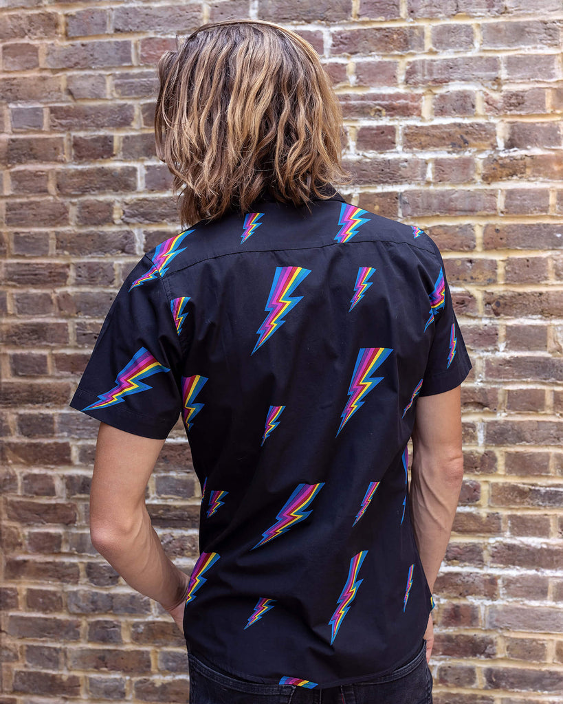 Rudi a male white tanned model with brown hair and a surfer style is wearing a black shirt with lightning bolt design against a brick wall.