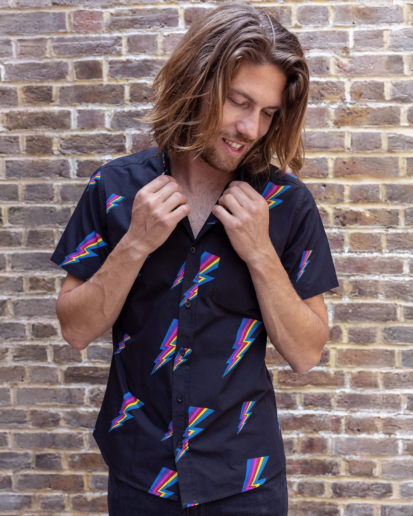 Rudi a male white tanned model with brown hair and a surfer style is wearing a black shirt with lightning bolt design against a brick wall
