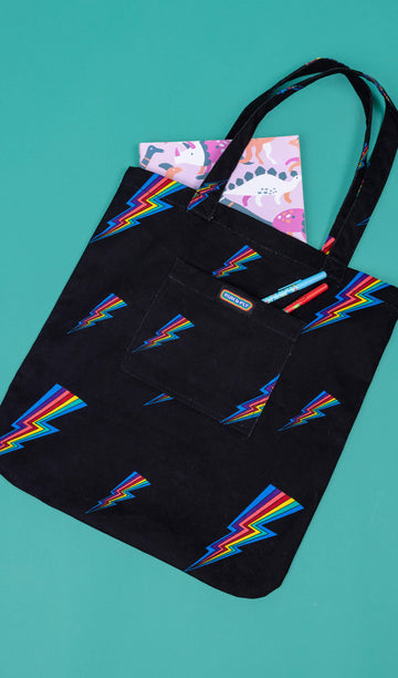 a black tote bag with all over rainbow lightning bold print. The tote bag is laid flat on a green background with a book in the main section of the bag the smaller front pocket has Run & Fly logo on the front.