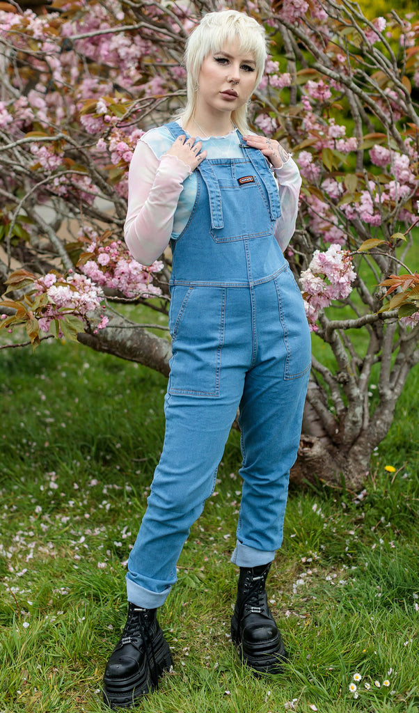 Flo a white blonde haired femme white model with tattoos is wearing light blue stone wash denim dungarees with a pastel long sleeve top and black platform trainers shot outside in front of a blossom tree in Shoreham, East Sussex by Amy Davies Photography. She is facing forward smising with both hands resting on the top roller straps.