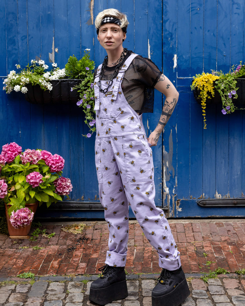 Jamie is wearing the Lavender bees stretch twill dungarees worn with a black fishnet top and platform boots, hem is rolled up. Model is posing toward the camera and standing in front of a garage with cobbled floors and flowers.