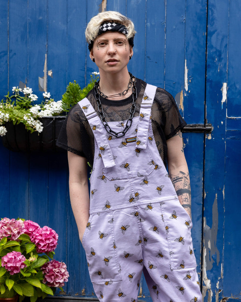 Jamie is wearing the Lavender bees stretch twill dungarees worn with a black fishnet top. Model is standing with hands in pockets, facing the camera and standing in front of a garage with cobbled floors and flowers.